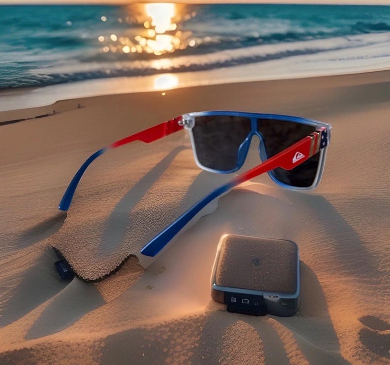 May include: Sunglasses with a red, white, and blue color scheme, resting on a sandy beach. The sunglasses have a rectangular frame and dark lenses. A small, rectangular, silver-colored device is also on the sand.