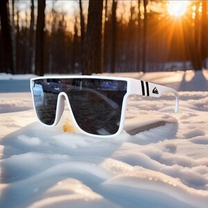 May include: White sunglasses with a wide frame and dark lenses. The sunglasses have black stripes and the Quiksilver logo on the temple. The sunglasses are resting on a snowy surface with a forest and sunlight in the background.