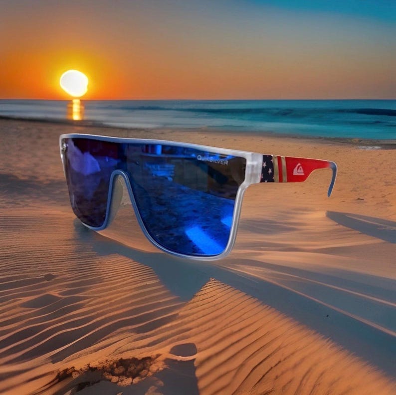 May include: Clear-framed sunglasses with blue lenses rest on a sandy beach. The temples feature a red, white, and blue design. The setting sun and ocean create a vibrant backdrop, reflecting in the lenses. The sunglasses are a stylish accessory.
