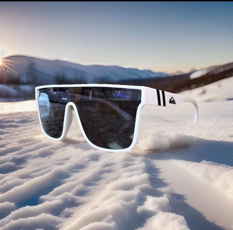 May include: White sunglasses with a wide frame and dark lenses. The sunglasses have the word "Quiksilver" printed on the lens. The arms of the sunglasses have black stripes and a logo. The sunglasses are resting on a snowy surface.