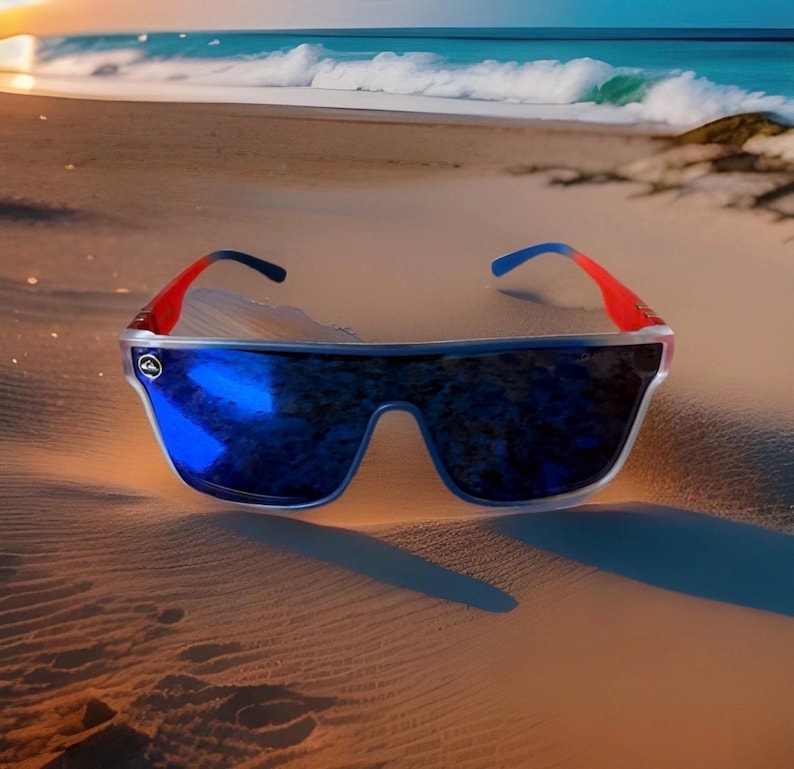 May include: Sunglasses with a wide, rectangular frame and mirrored blue lenses. The temples are red and blue, resting on a sandy beach with the ocean in the background. The sunglasses are designed for outdoor use.