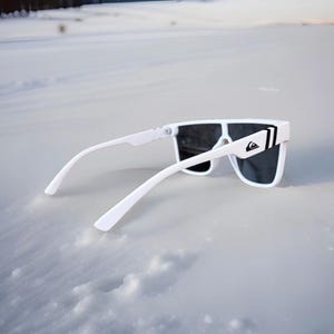 May include: White sunglasses with dark lenses and a white frame. The sunglasses have a rectangular shape and a logo on the side. The sunglasses are resting on a snowy surface.