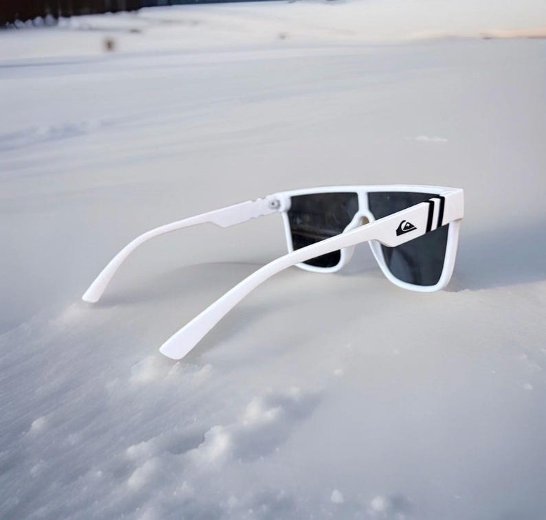 May include: White sunglasses with dark lenses and a white frame. The sunglasses have a rectangular shape and a logo on the side. The sunglasses are resting on a snowy surface.