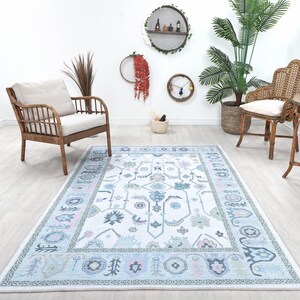 May include: A rectangular area rug with a light blue and white floral pattern. The rug has a border with a repeating design and is surrounded by wicker furniture, plants, and decorative wall hangings. The room has light wood flooring.