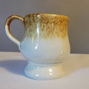 May include: A white ceramic mug with a brown glaze on the top half. The mug has a rounded body and a flared base.