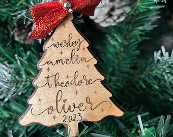 May include: A wooden Christmas tree ornament with the names "Wesley", "Amelia", "Theodore", and "Oliver" engraved on it. The ornament also includes the year "2023" and a red glitter bow.