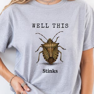 Puede incluir: Una camiseta gris claro con una ilustración detallada de una chinche marrón centrada en el pecho. Sobre el insecto, el texto dice WELL THIS en negro, y debajo del insecto, el texto dice STINKS en negro, usando una fuente estilo máquina de escribir.