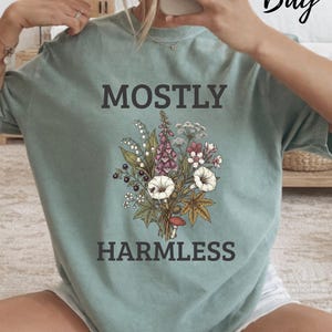 Peut inclure: Un t-shirt à manches courtes vert sauge orné d'une illustration botanique composée de fleurs sauvages, de baies et d'un petit champignon. Le texte MOSTLY HARMLESS est imprimé en lettres capitales gris foncé au-dessus et en dessous du motif floral.