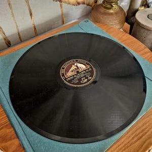 Pode incluir: Um disco de vinil preto vintage com o rótulo "HIS MASTER'S VOICE". O disco está colocado dentro de uma capa azul sobre uma mesa de madeira. O rótulo apresenta um cão a ouvir um gramofone. Um objeto de coleção de música clássica.