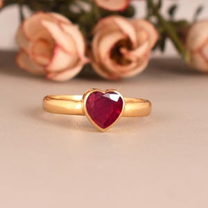 以下が含まれることがあります： ハート型の深紅色の宝石が付いたゴールドリング。リングは、背景にぼやけた花がある、柔らかくニュートラルな背景にセットされています。宝石の幅は約1.3cmです。