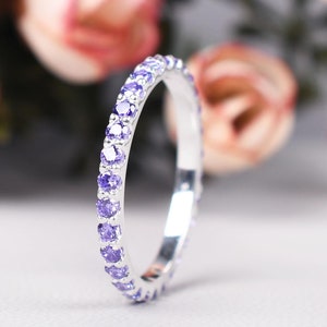 May include: A silver ring with a band of small, round, purple gemstones. The ring is in focus against a blurred background of pink and green flowers. The ring is a delicate piece of jewelry.