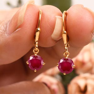 May include: A pair of gold drop earrings featuring red ruby gemstones. The earrings have a simple design with a small round ruby suspended from a fine gold chain.
