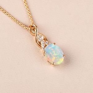 May include: A gold-colored necklace with a pendant featuring an oval opal gemstone. The pendant is accented with small, clear gemstones and has a decorative, intertwined design. The chain is a delicate, linked style.