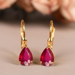 May include: A pair of gold-coloured leverback earrings, each with a teardrop-shaped, faceted, deep red gemstone. The earrings are set against a neutral background, with blurred pink flowers in the background.