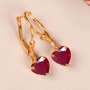 May include: A pair of gold-coloured leverback earrings, each with a heart-shaped, deep red gemstone. The earrings are set against a soft, neutral background, with floral accents in the upper corners.