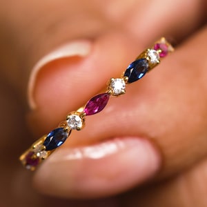 Könnte beinhalten: Ein goldener Ring mit abwechselnd geschliffenen blauen Saphiren, Rubinen und runden Diamanten. Der Ring wird in einer Hand gehalten, wodurch das filigrane Design und die leuchtenden Farben der Edelsteine hervorgehoben werden.