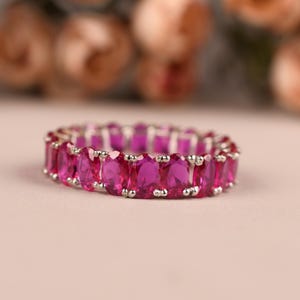 May include: A silver ring featuring a continuous band of oval-cut, bright pink gemstones. The ring is set on a pale pink surface, with blurred peach-coloured flowers in the background. The gemstones are closely set, forming a full eternity band.