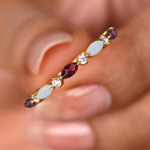 Może przedstawiać: Delikatny złoty pierścionek z naprzemiennymi kamieniami szlachetnymi. Pierścionek prezentuje owalne opale, ciemnoczerwone granaty i małe, okrągłe diamenty. Kamienie szlachetne osadzone są w cienkiej, złotej oprawie, tworząc wyrafinowany i elegancki design.