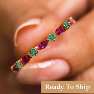 May include: A gold ring with alternating ruby and emerald gemstones. The ring features marquise-cut rubies and square-cut emeralds set in a delicate band. The ring is shown against a blurred background. Text on the image reads "Ready To Ship".