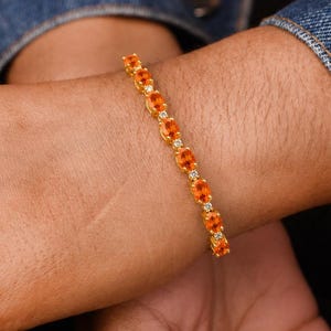 Può includere: Un bracciale dorato con pietre preziose ovali arancioni e piccole pietre trasparenti. Il bracciale è indossato su un polso, con una giacca di jeans sullo sfondo. È un gioiello.