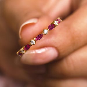 Könnte beinhalten: Ein goldener Ring mit abwechselnd tropfenförmigen roten Edelsteinen und runden, klaren Edelsteinen. Der Ring wird in einer Hand gehalten, wodurch das filigrane Design und der Glanz des Schmucks hervorgehoben werden.