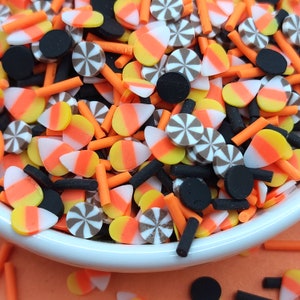 May include: A close-up of a white bowl filled with Halloween-themed polymer clay sprinkles. The sprinkles include candy corn, orange sticks, black cylinders, and brown and white pinwheel shapes. The background is orange.