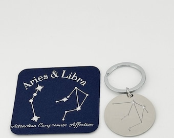 May include: A dark blue rectangular card featuring the text Aries & Libra and Attraction Compromise Affection with white constellation graphics, alongside a circular silver metal keychain engraved with the Libra constellation.