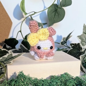 May include: A handmade crochet doll in the shape of a character with a pink hood, ears, and a yellow bow. The doll has a white face with black eyes and a yellow beak. The doll is sitting on a small wooden box, surrounded by greenery.