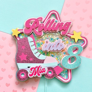 May include: A pink and blue roller skate cake topper with glitter and a star shape. The topper says "Rolling into 8" and "Mia".