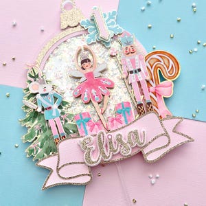 May include: A pink and gold glitter cake topper featuring a ballerina, nutcracker, and mouse from The Nutcracker ballet. The topper includes a banner with the name "Elisa" and a number 1, perfect for a birthday celebration.