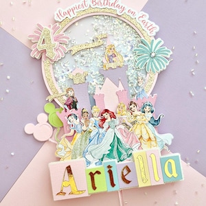 May include: A multi-layered cake topper with a princess theme. The topper features a castle, the number 4, and the name "Ariella" spelled out in colorful blocks. The words "Happiest Birthday on Earth" are written in a circular design.