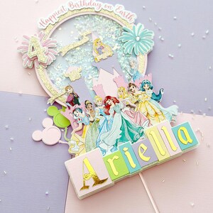 May include: A multi-layered birthday cake topper with a princess theme. The topper features a glittery gold and pink circular frame with the text "Happiest Birthday on Earth". The number "4" is displayed, along with the name "Ariella".
