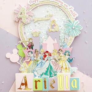 May include: A multi-layered birthday cake topper featuring Disney princesses, a castle, and the number 4. The topper includes the text "Happiest Birthday on Earth" and the name "Ariella" in colorful blocks. Glitter and floral accents complete the design.