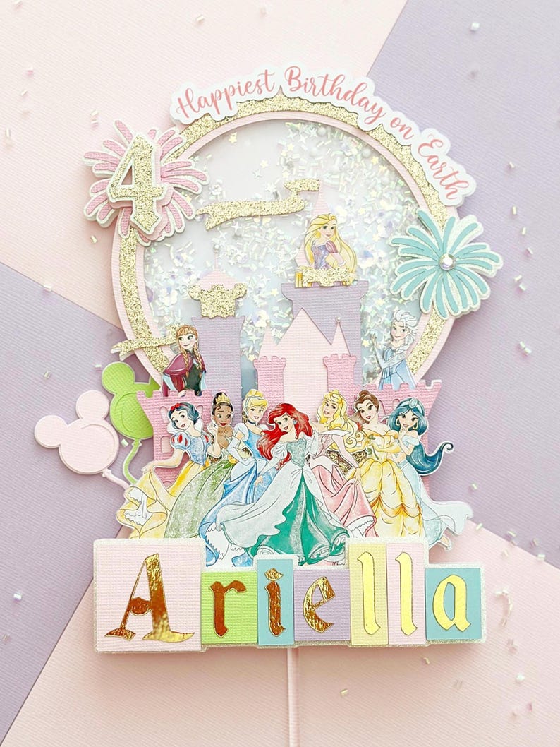 May include: A multi-layered birthday cake topper featuring Disney princesses, a castle, and the number 4. The topper includes the text "Happiest Birthday on Earth" and the name "Ariella" in colorful blocks. Glitter and floral accents complete the design.