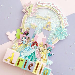 May include: A multi-layered birthday cake topper featuring Disney princesses. The topper includes the name "Ariella" in colorful block letters, a castle, and the phrase "Happiest Birthday on Earth". The number "4" is also included.