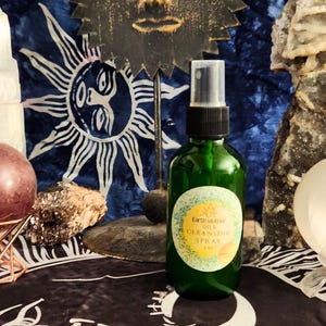 May include: A green glass spray bottle with a label that reads "Earth Mother Oils Cleansing Spray" sits on a black cloth. A decorative sun face and a blue patterned background are visible. The bottle is approximately 5 inches tall.