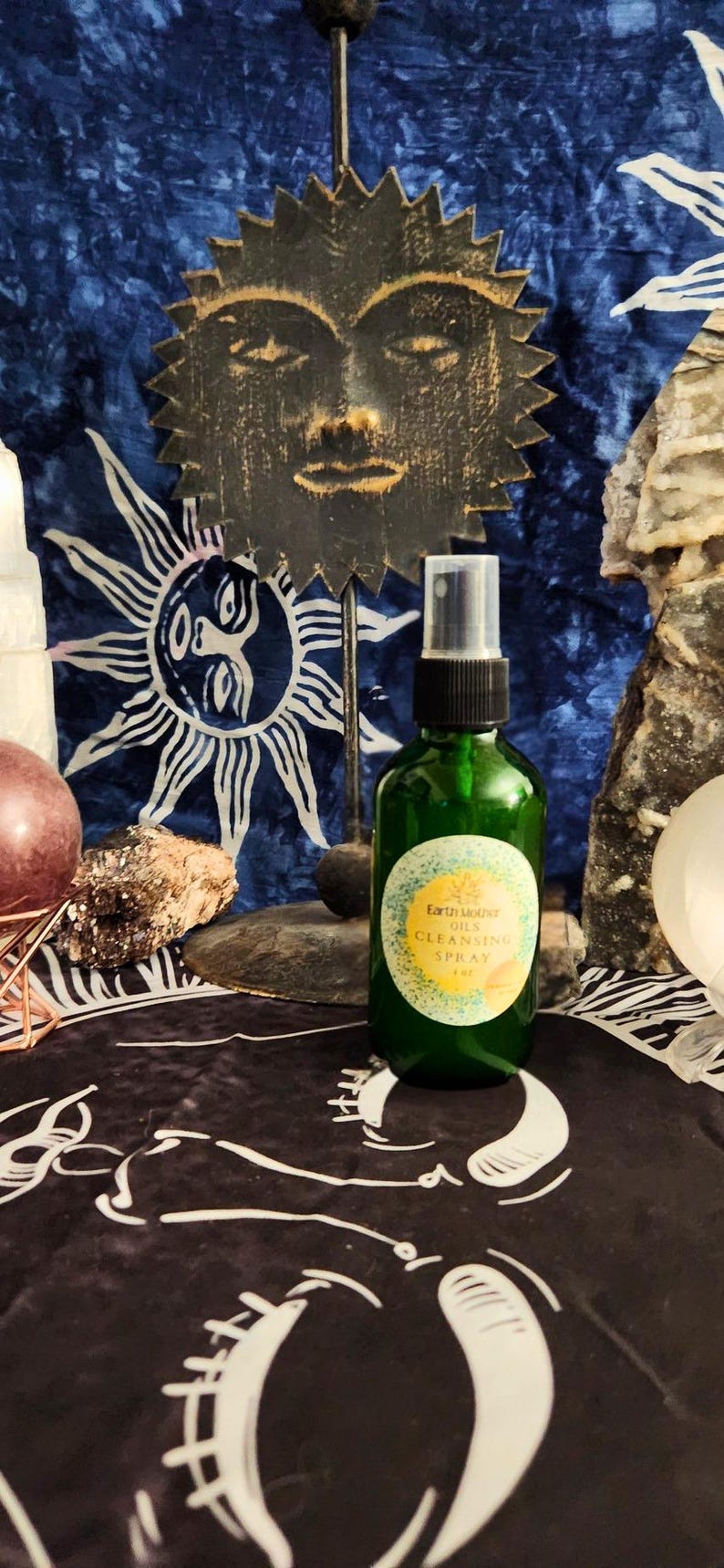 May include: A green glass spray bottle with a label that reads "Earth Mother Oils Cleansing Spray" sits on a black cloth. A decorative sun face and a blue patterned background are visible. The bottle is approximately 5 inches tall.