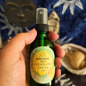 May include: A green glass spray bottle labeled "Earth Mother Oils Cleansing Spray" with a yellow and green label. The bottle contains 2 fluid ounces of product. The background is a dark blue fabric with sun and star designs.