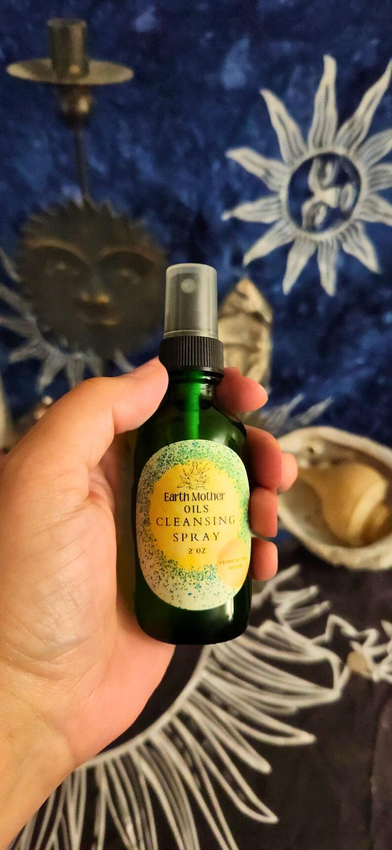 May include: A green glass spray bottle labeled "Earth Mother Oils Cleansing Spray" with a yellow and green label. The bottle contains 2 fluid ounces of product. The background is a dark blue fabric with sun and star designs.
