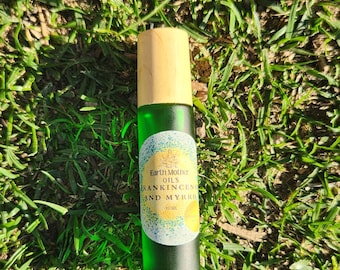 May include: A green glass essential oil bottle with a wooden cap. The label reads "Earth Mother Oils Frankincense and Myrrh". The bottle is set against a backdrop of green grass.