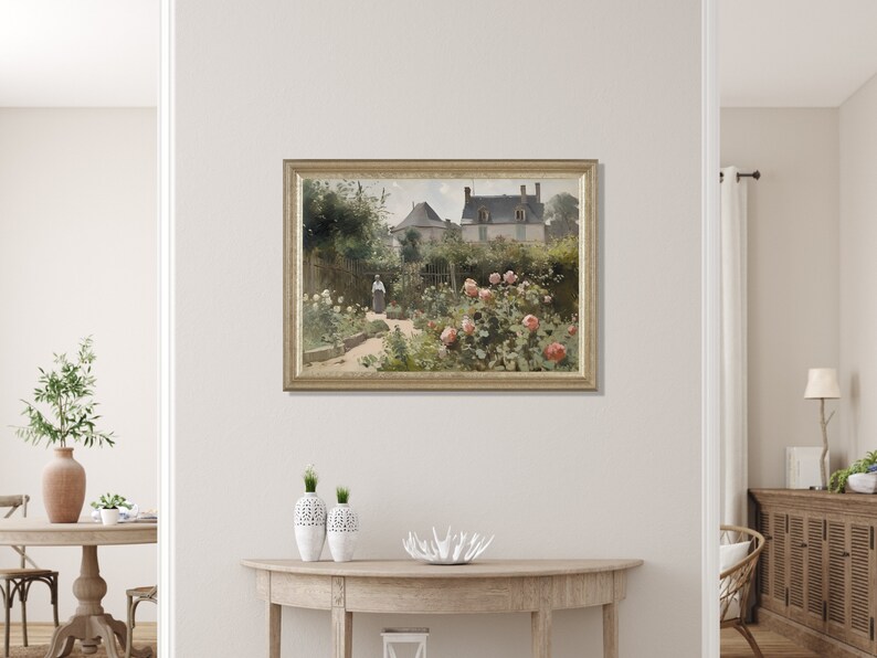 May include: A framed painting depicts a garden scene with a house in the background. The garden features a path, lush greenery, and blooming pink roses. A person in a long dress is walking in the garden. The painting is in a gold frame.