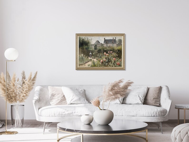 May include: A framed painting depicts a garden scene with a house in the background. The garden features rose bushes with pink blooms, a pathway, and figures. The painting is displayed on a white wall above a gray sofa, enhancing the room's decor.