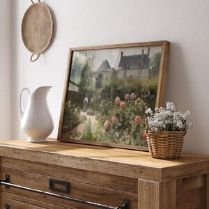May include: A framed landscape painting of a garden with a house in the background. The painting is in a wooden frame and is displayed on a wooden dresser. A white pitcher and a small basket of white flowers are also on the dresser.