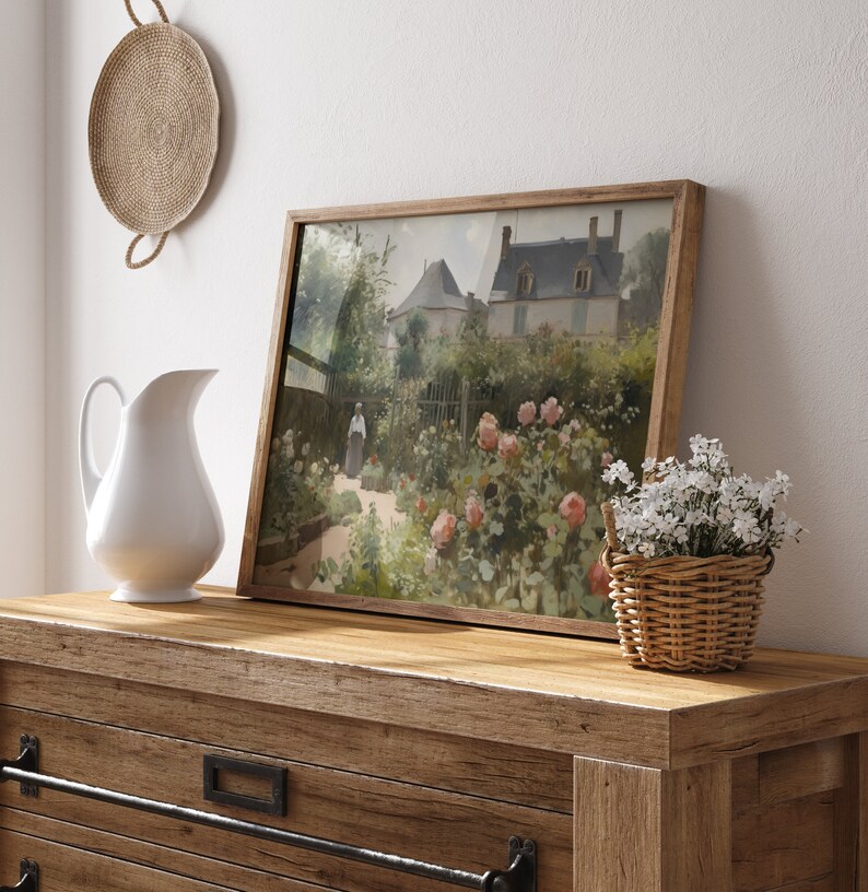 May include: A framed landscape painting of a garden with a house in the background. The painting is in a wooden frame and is displayed on a wooden dresser. A white pitcher and a small basket of white flowers are also on the dresser.
