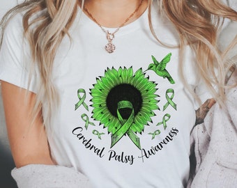 Peut inclure: T-shirt blanc avec un motif de tournesol vert, des rubans verts et un colibri. Le texte "Cerebral Palsy Awareness" est imprimé en noir. Le t-shirt est à col rond.