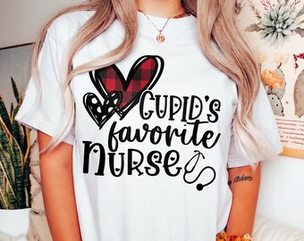 以下が含まれることがあります： 赤いチェック柄のハートと「Cupid's favorite nurse」というテキストと聴診器のグラフィックが描かれた白いTシャツです。