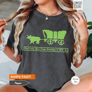Könnte beinhalten: Dunkelgraues T-Shirt mit einem pixeligen grünen Planwagen und Ochsen-Grafik. Der Text "You have died from listening to RFK Jr." steht darunter. Unisex-Größe.