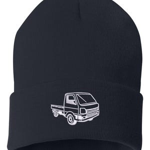 May include: A dark navy blue knit beanie featuring a white embroidered graphic of a small pickup truck on the front cuff. The hat has a ribbed texture and a folded brim, showcasing a detailed outline of the vehicle with visible wheels and cab.