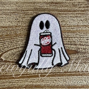May include: Embroidered patch featuring a white ghost with black eyes holding a Dr. Pepper can. The patch has a black border and is on a wooden background. The text "Sewing Hands" is visible at the bottom.
