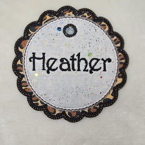 May include: A round, white patch with the name "Heather" embroidered in black script. The patch has a scalloped edge with a leopard print design and is embellished with glitter.